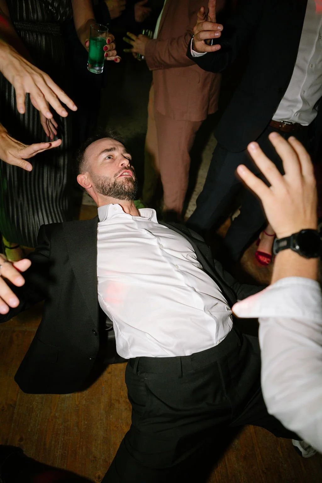 A man in a tuxedo lying on the ground with a distressed expression as several people surround him at a party or social gathering.