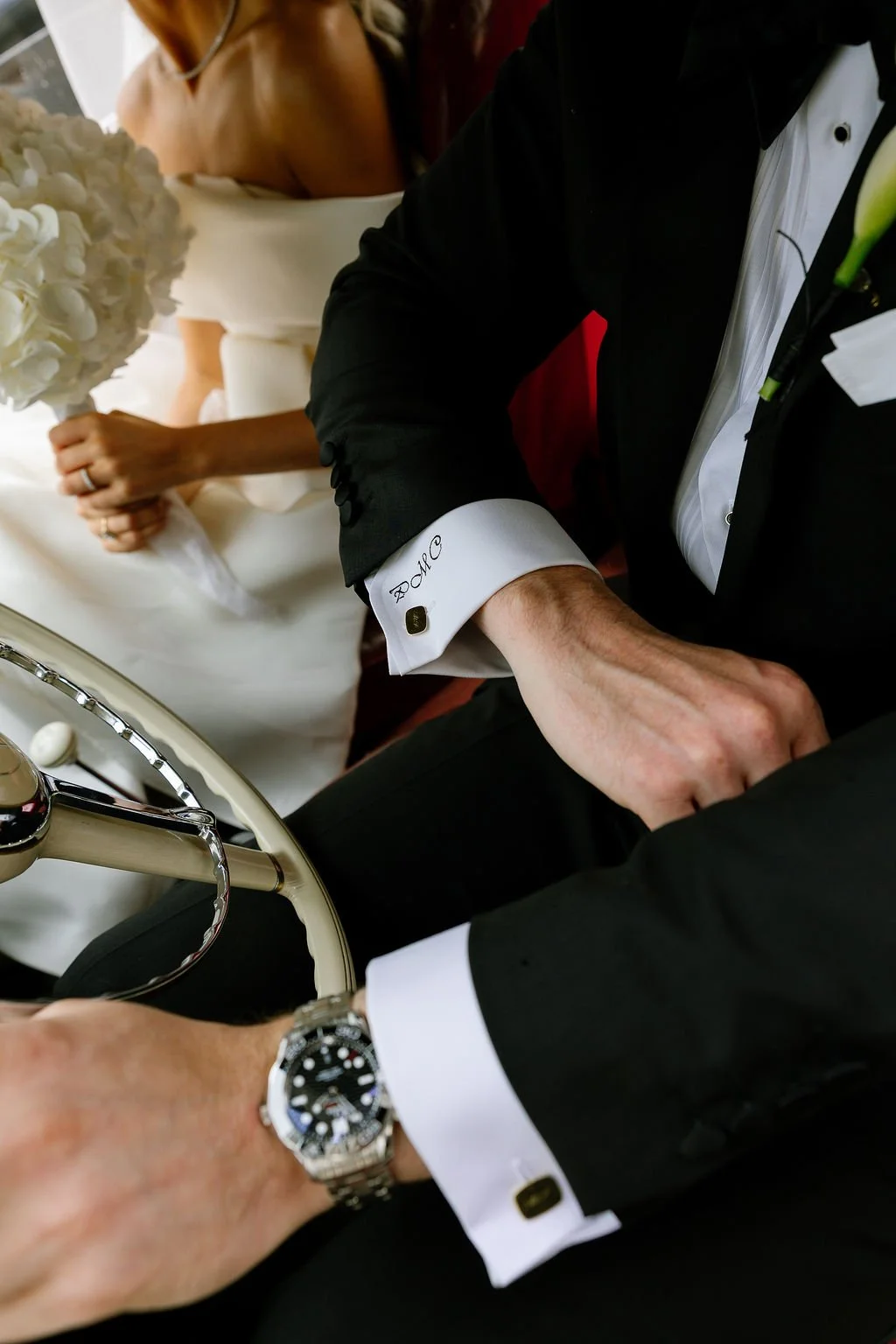 Close-up of two men's hands in tuxedos, one wearing a Rolex watch, the other resting on a vintage steering wheel, with a bride in a white wedding dress holding a bouquet of white flowers in the background.