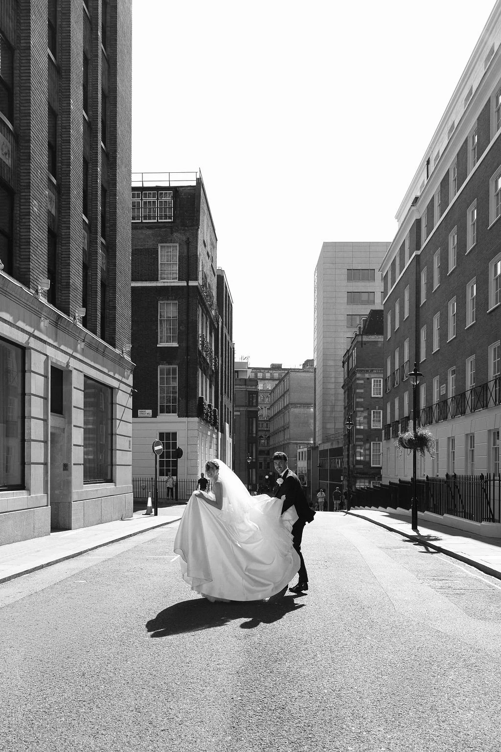 A bride in a wedding dress and veil being lifted by a groom in a suit on a city street with tall buildings, illuminated by sunlight, in black and white.