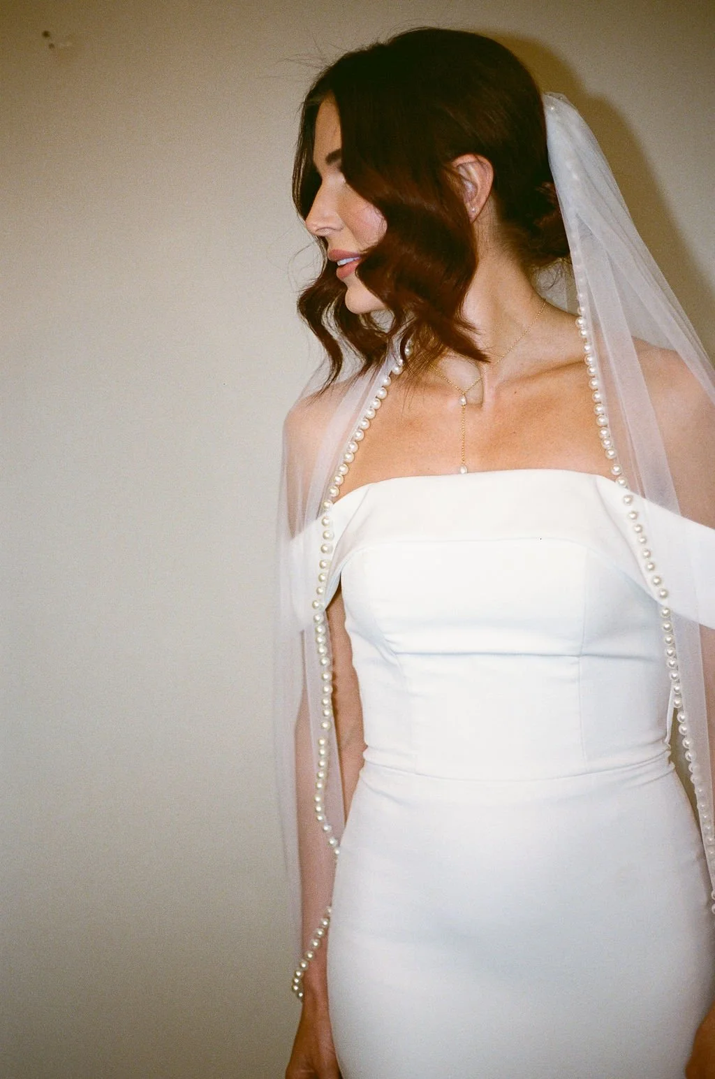 A woman with shoulder-length wavy brunette hair wearing a white strapless wedding dress and a pearl-embellished veil, looking to the left against a plain background.