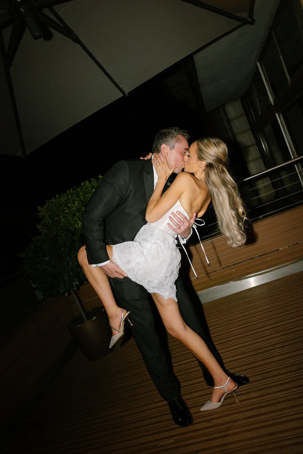 A man in a black suit and a woman in a white dress are sharing a kiss, with the woman lifted by the man at night on a wooden deck outside.