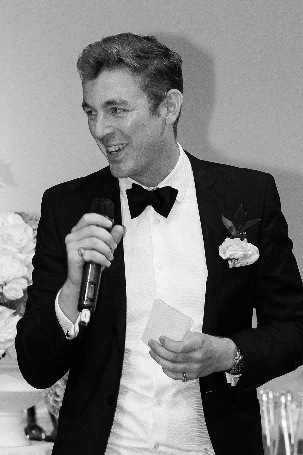 A man in a tuxedo with a bow tie, holding a microphone and a small notepad, smiling at a formal event.