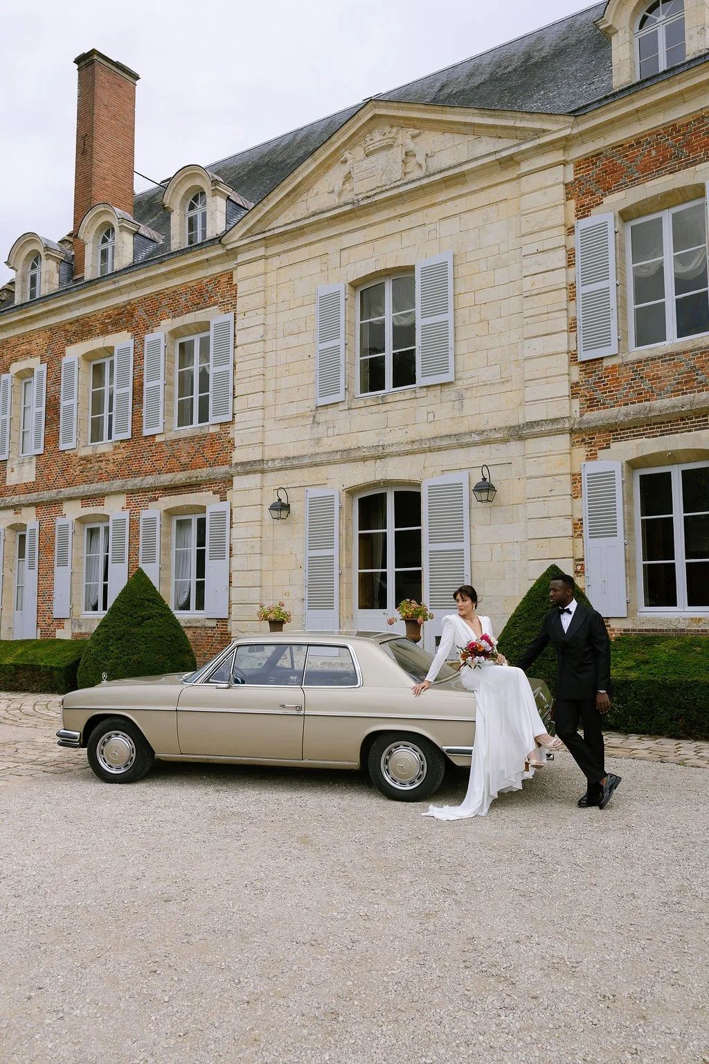A bride and groom pose next to a vintage Mercedes-Benz car in front of an elegant, historic building with large windows and white shutters. The bride is seated on the car, holding a bouquet, and the groom stands beside her, holding her hand. The scen