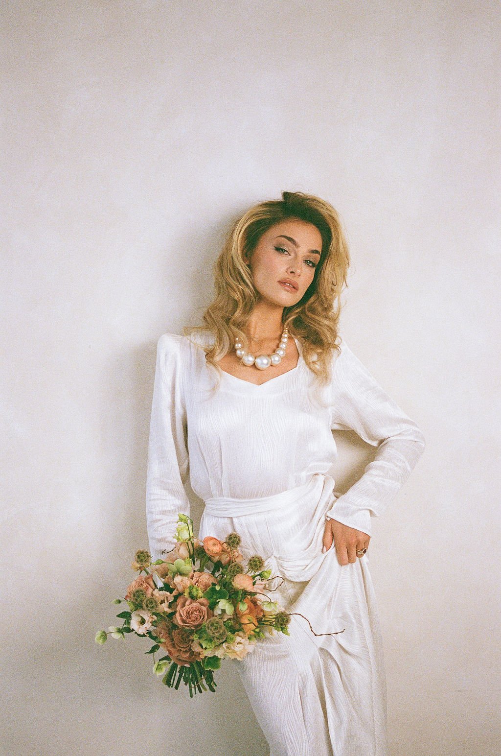 A woman with blonde hair wearing a white dress and pearl necklace, holding a bouquet of pink and green flowers, standing against a plain light-colored wall.