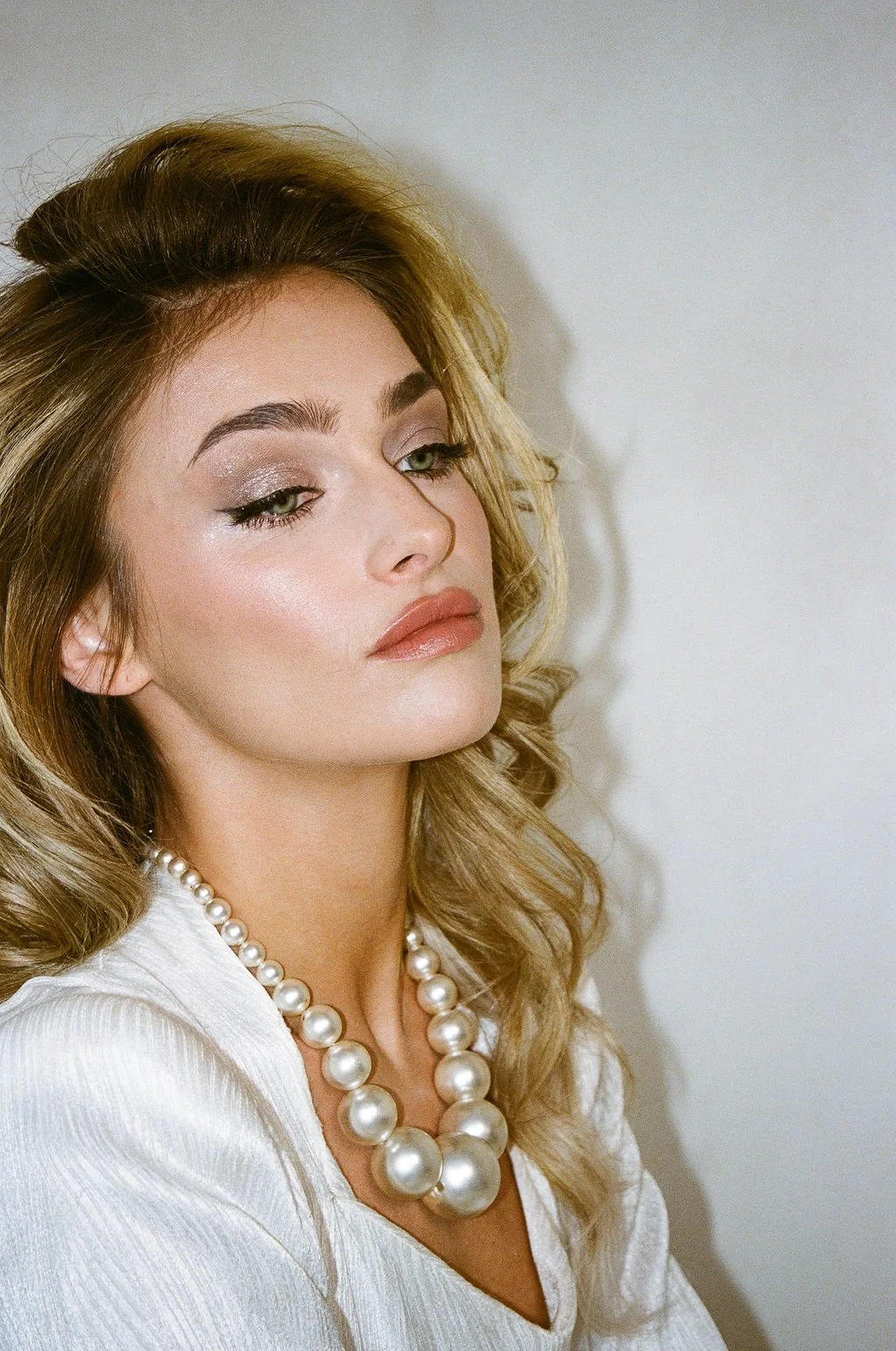 A woman with blonde, wavy hair, wearing pearl necklace and a white top, posing with a neutral expression against a plain light background.