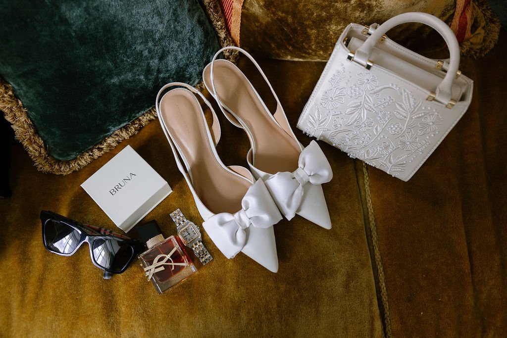 White high-heeled shoes with large bows on the front, a white handbag with floral embroidery, black sunglasses, a small perfume bottle, a box labeled BRUNA, and a watch, all placed on a brown surface with pillows in the background.