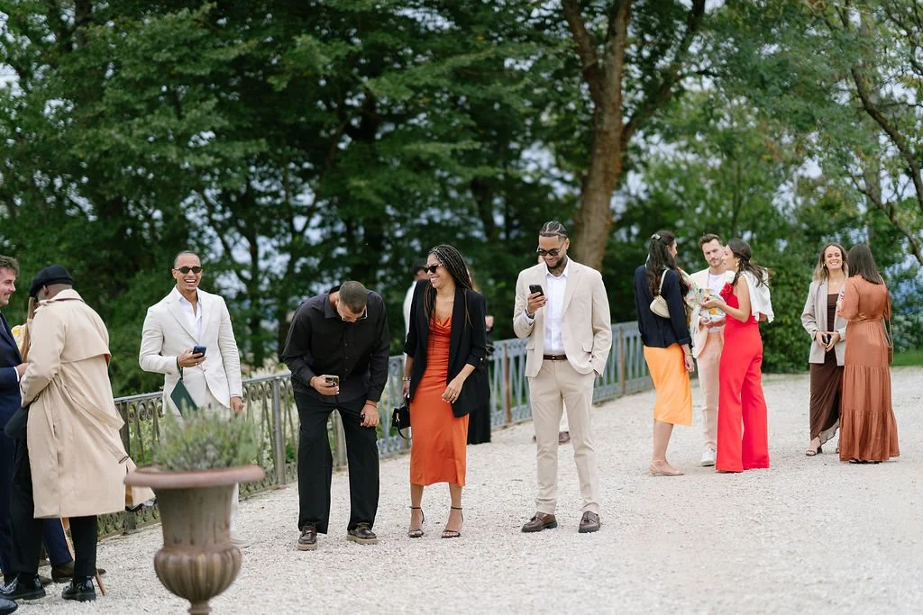 A group of people in stylish clothing standing outdoors on a gravel path, some looking at their phones, with trees in the background.