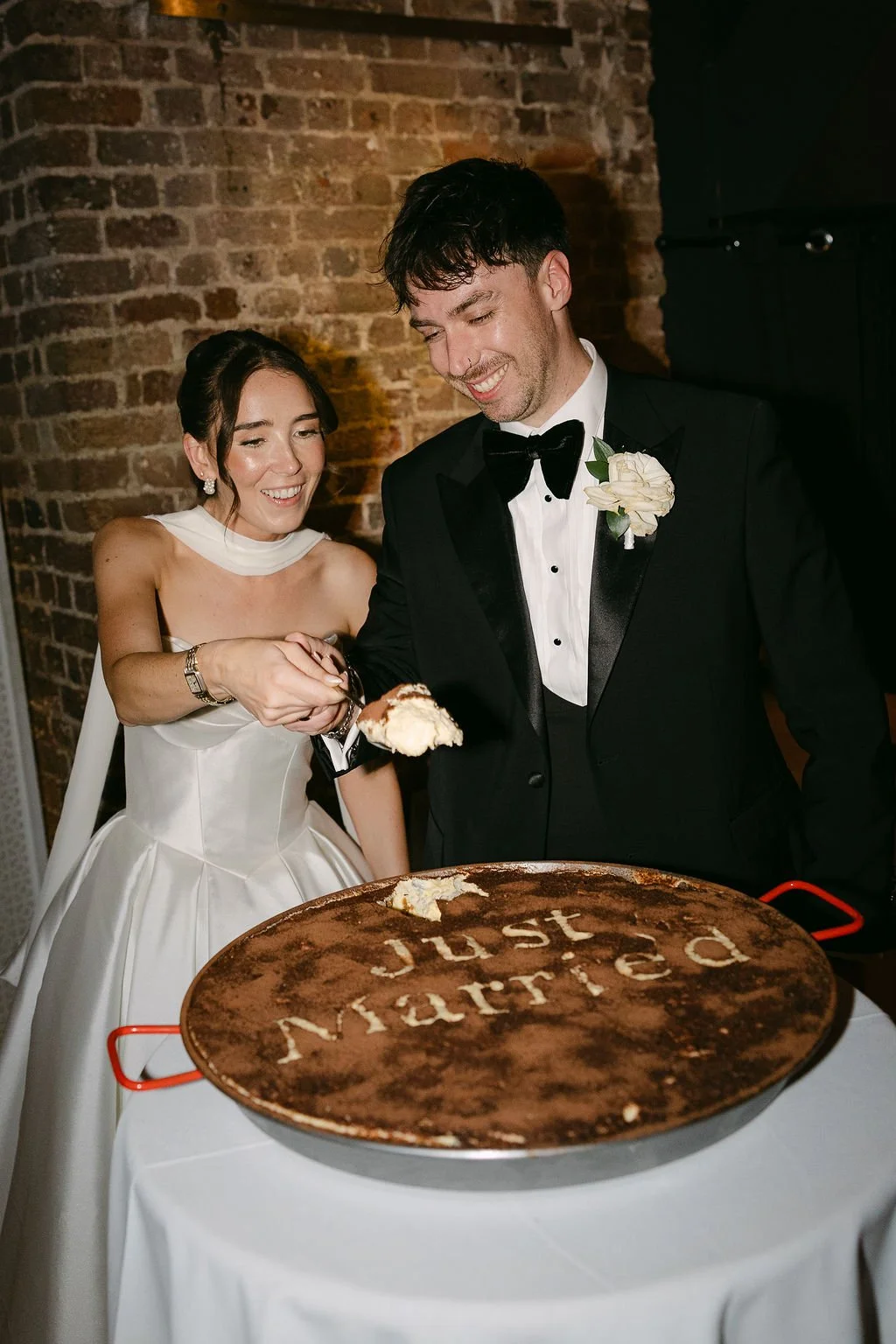 A bride and groom cutting a wedding cake with the words 'just married' on it, standing against a brick wall.
