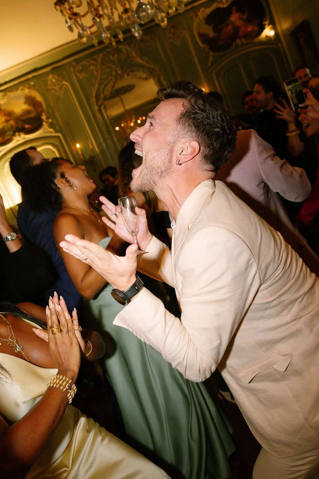 A man with dark hair and a beard, wearing a light-colored suit, is laughing and holding a champagne flute at a lively celebration in an ornate, elegant room with chandeliers and intricate wall decorations.