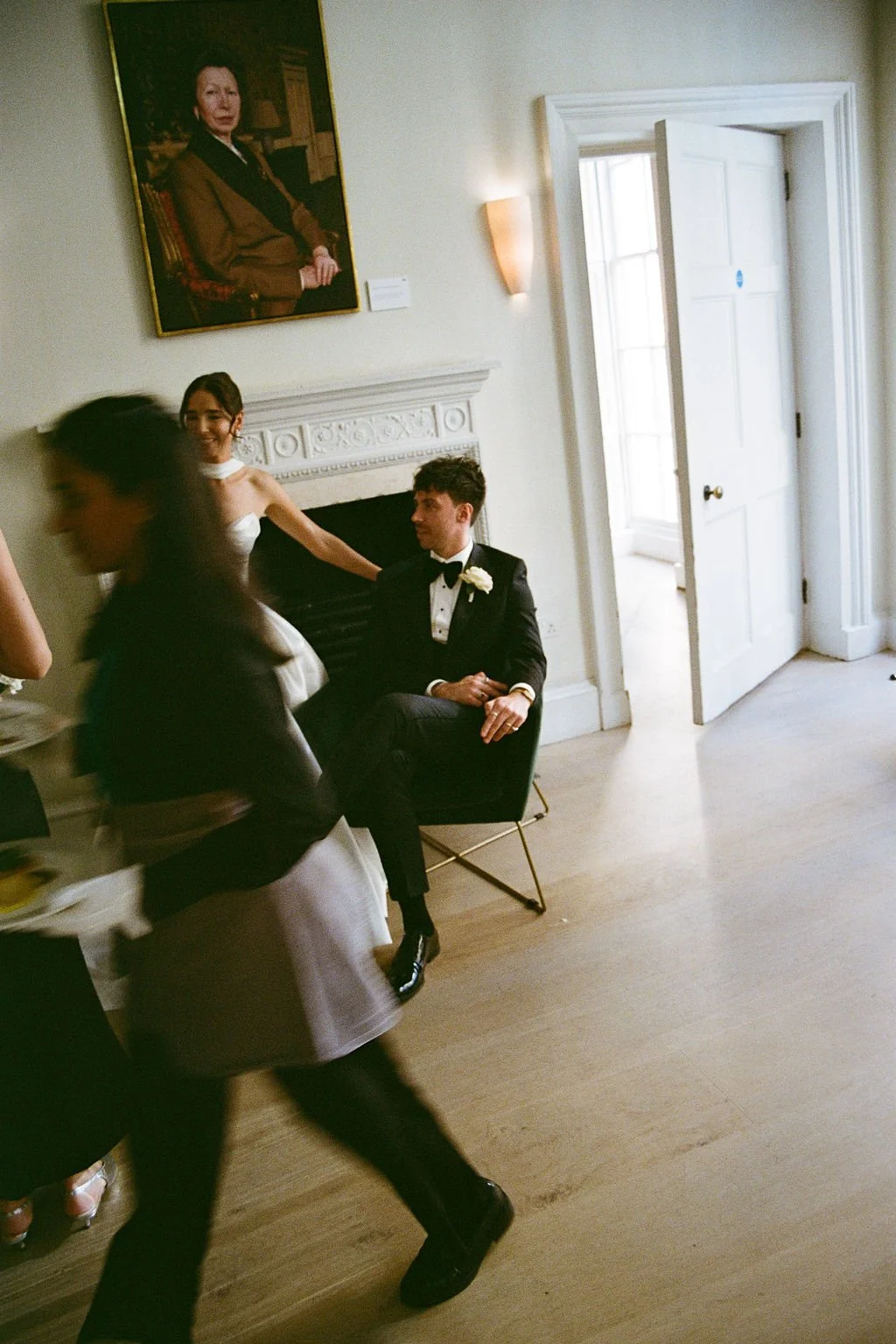 A man in a tuxedo sitting in a room, with a woman in a white dress and other people visible, a portrait of a woman on the wall, and an open door letting in light.