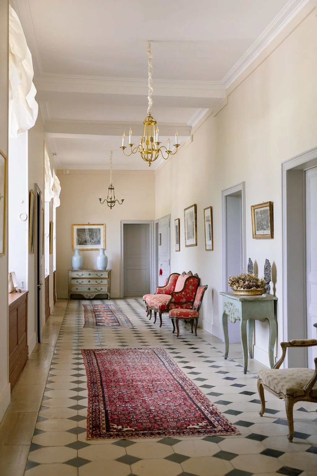Elegant hallway with checkerboard tile floor, red patterned rugs, vintage chairs, framed artwork, and antique furniture.