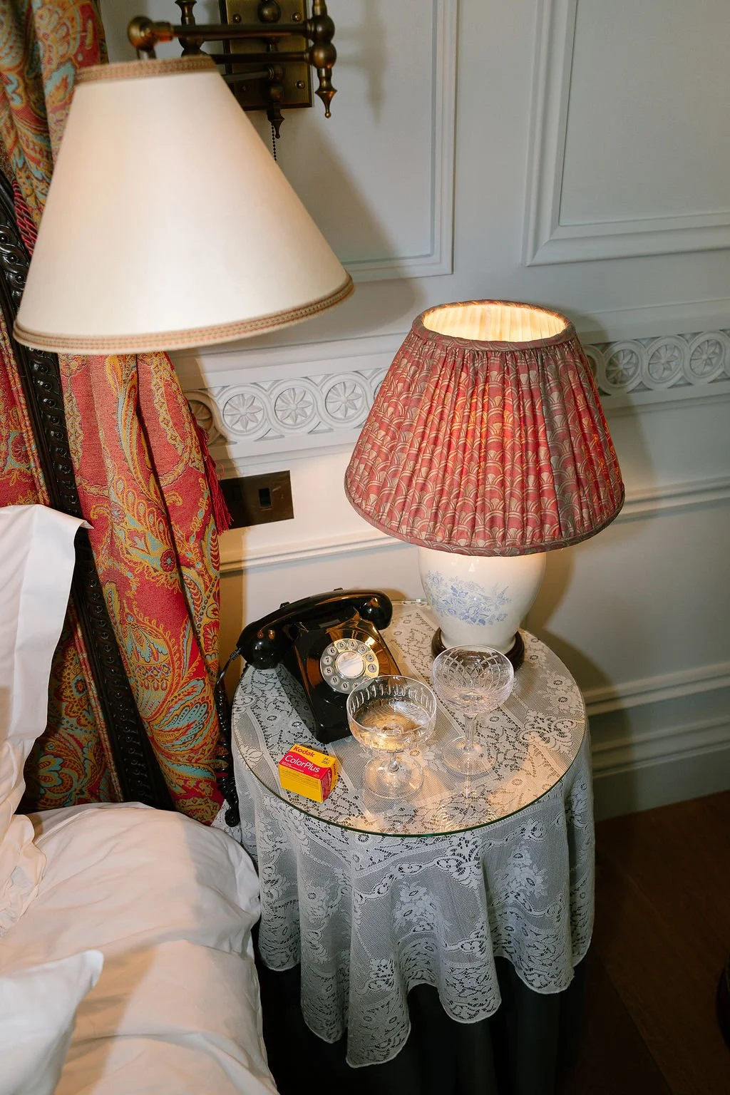A nightstand with a vintage black rotary phone, a box of Kodak ColorPlus 200 film, and two empty glasses. A table lamp with a floral-patterned shade is plugged into the wall, which also features ornate white molding and patterned drapes nearby.