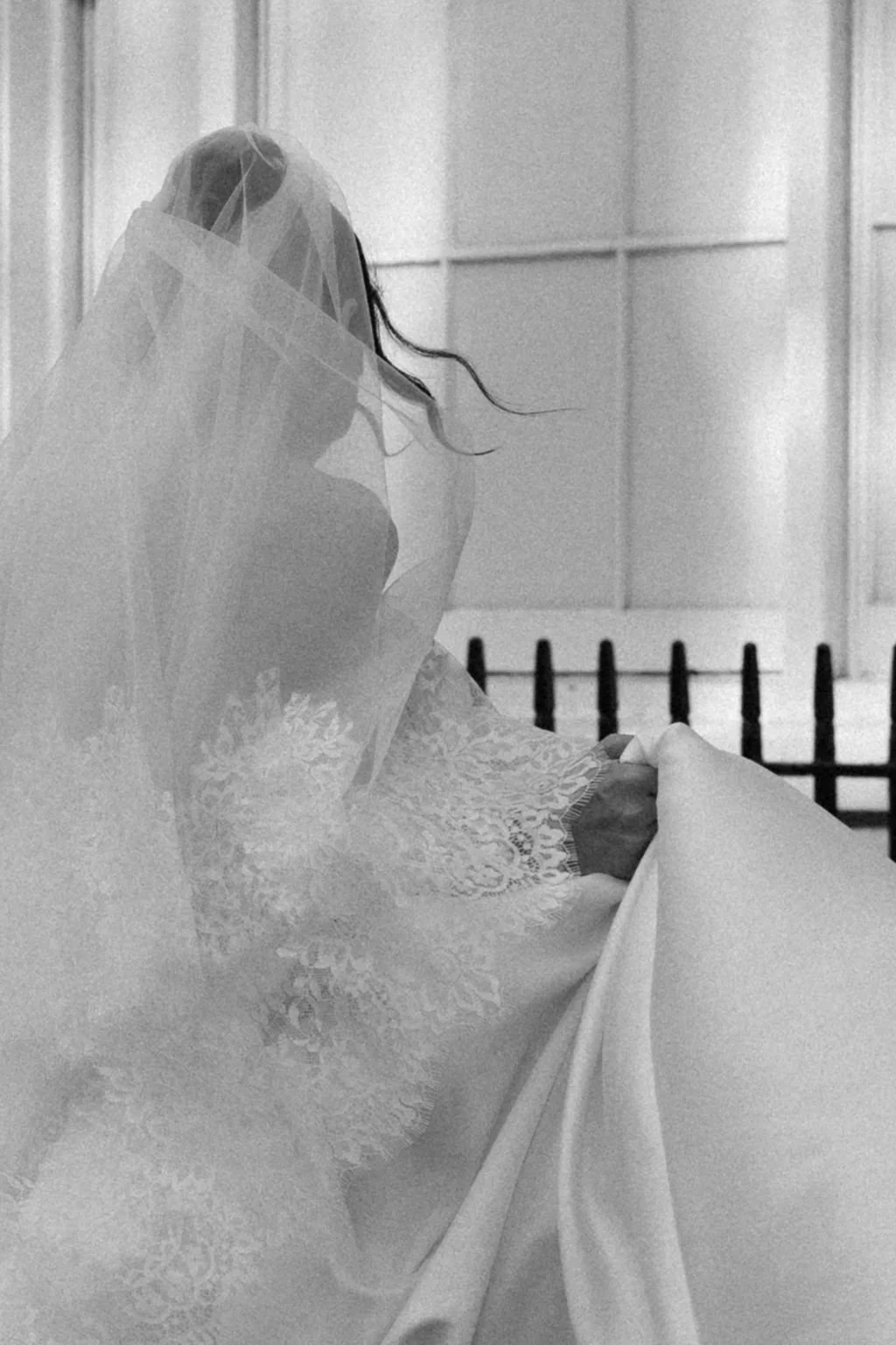 A bride in a wedding dress and veil holding her train, inside a building with light-colored walls.