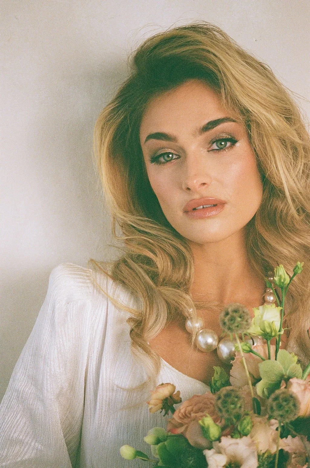 A woman with blonde wavy hair, blue eyes, wearing a white top and pearls, holding a bouquet of flowers.