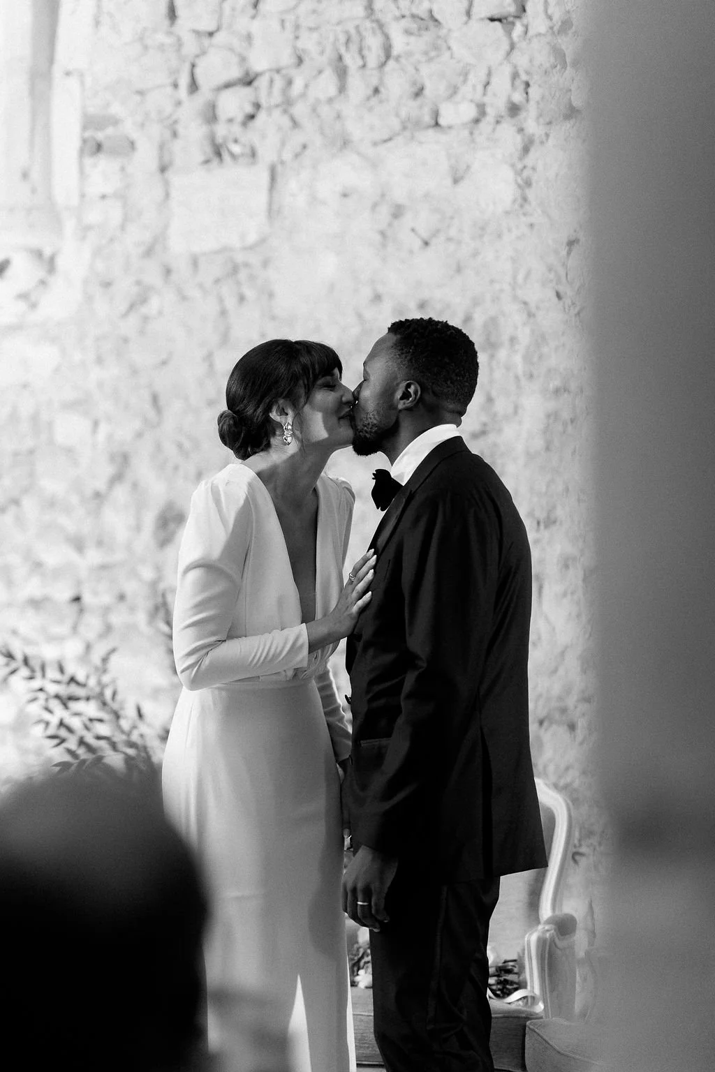 A black and white photograph of a bride and groom sharing a kiss at their wedding ceremony, standing against a textured brick wall.