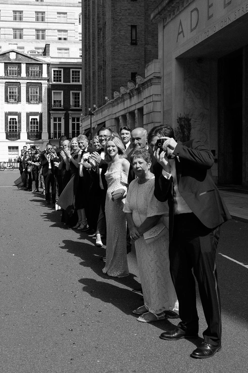 A line of wedding guests standing on a London street, smiling and taking photos.