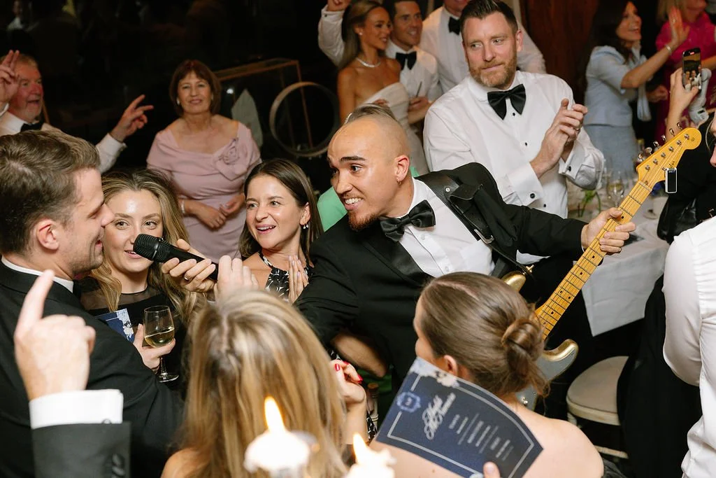 People at a wedding reception, some dressed in formal attire, with a band playing live music and guests dancing and singing.