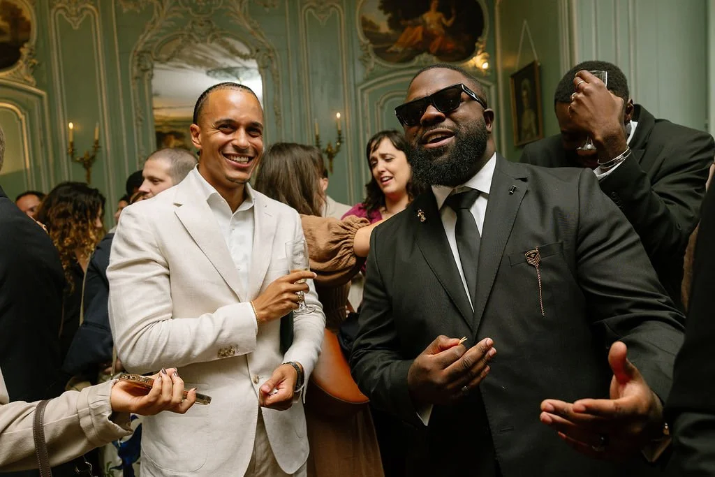 Group of people at a party, smiling and enjoying drinks in an elegant room with ornate decor and paintings.