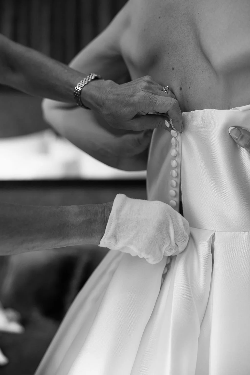 Close-up of an older person helping a bride button her wedding dress, showing their hands and part of the dress in black and white.