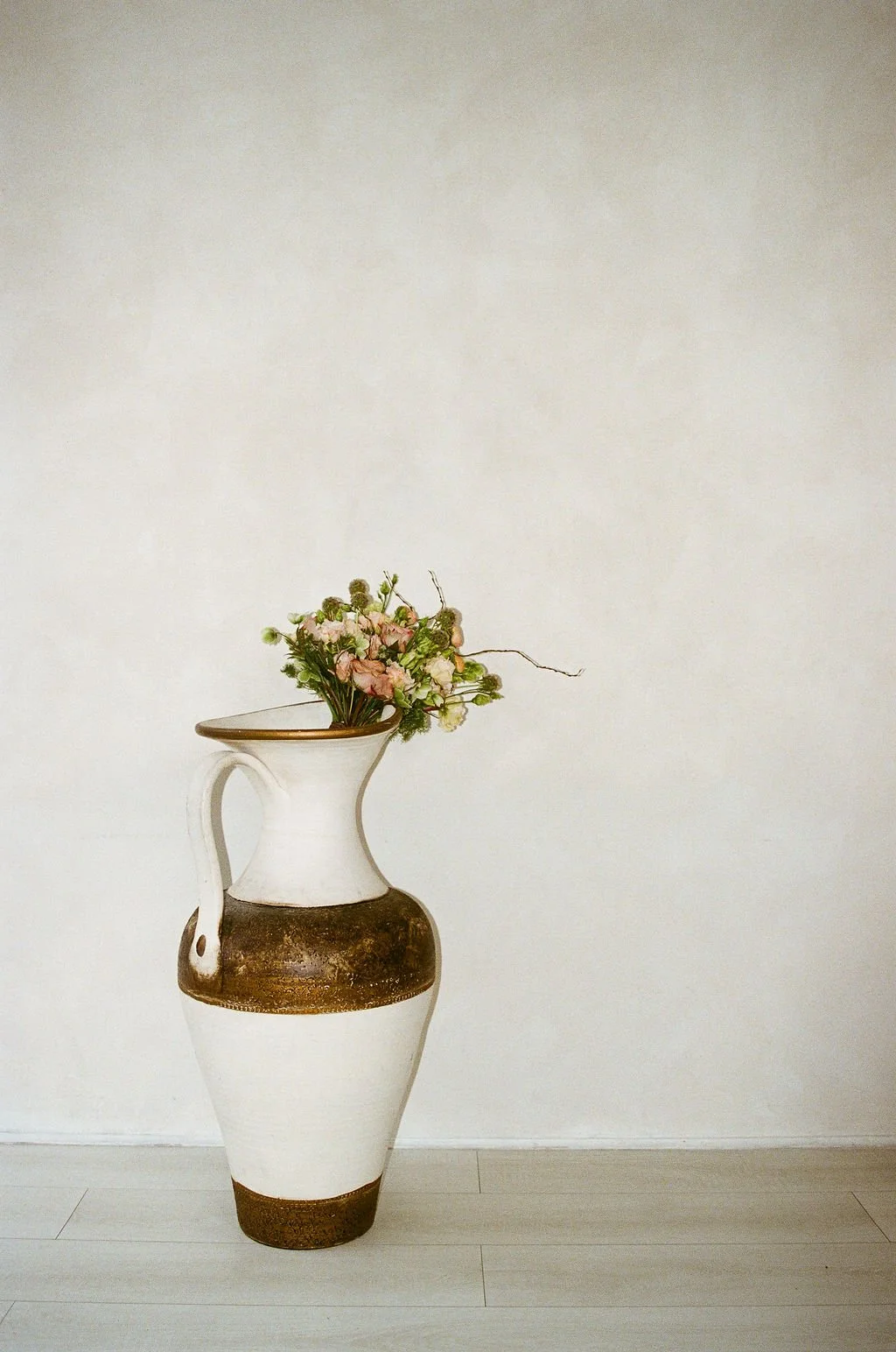 A tall, white ceramic vase with a brown band around its middle, placed against a plain white wall on a light-colored wooden floor, holding a bouquet of pink and white flowers with green foliage.