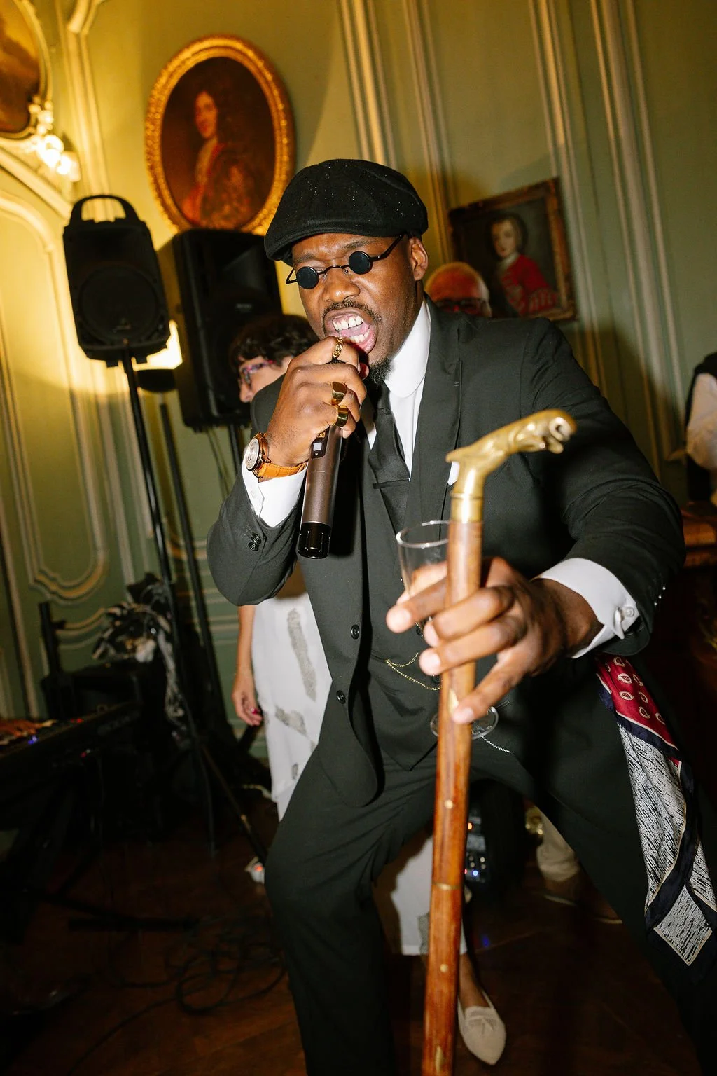 A man dressed in a black suit, white shirt, black tie, and black cap, with round sunglasses, holding a microphone in one hand and a glass of wine in the other, performing at a party or event in a room with ornate green walls, framed portraits, and so