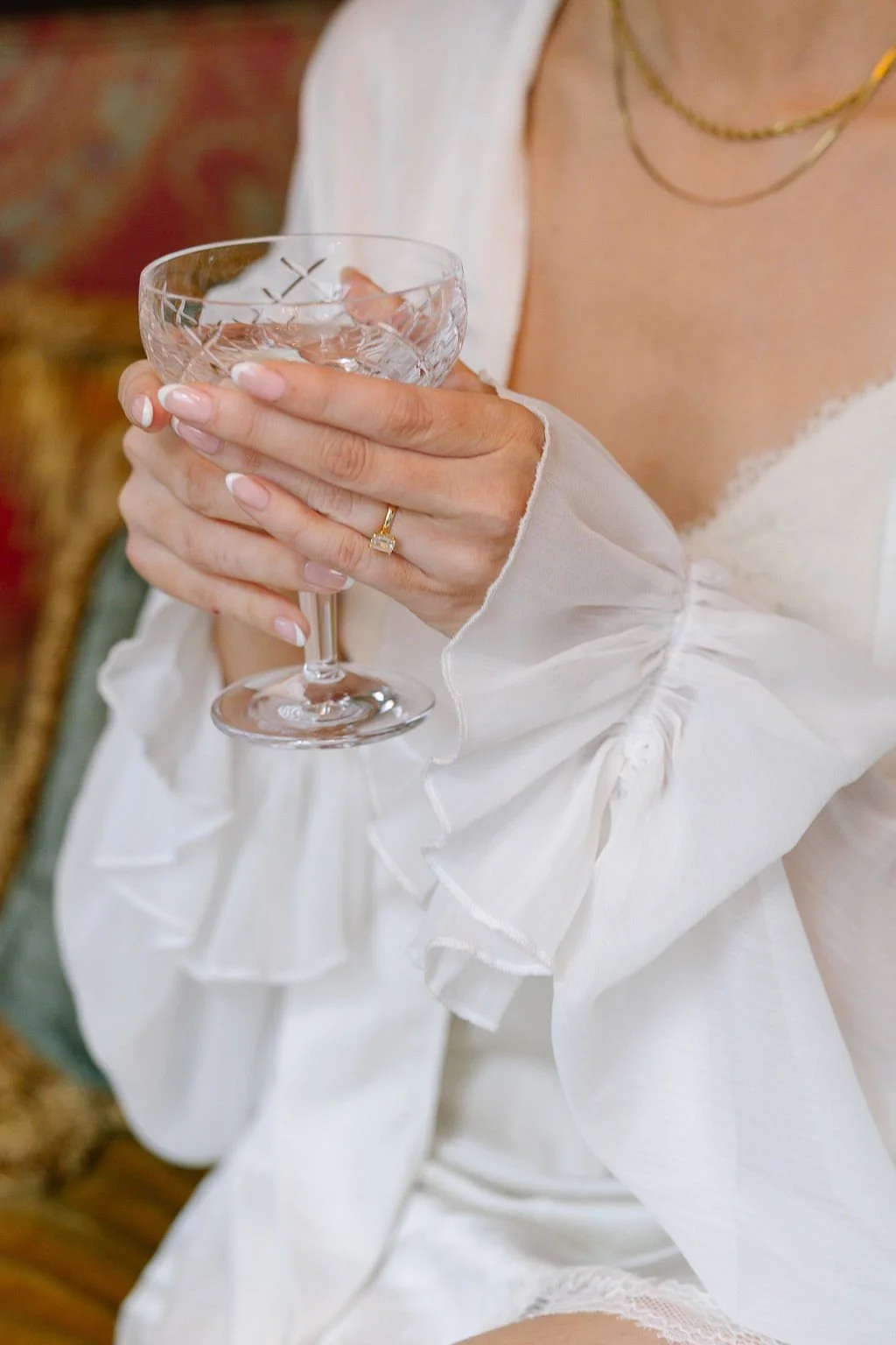 A woman in a white dress with ruffled cuffs holding a crystal glass with a drink, wearing a gold ring and necklace.