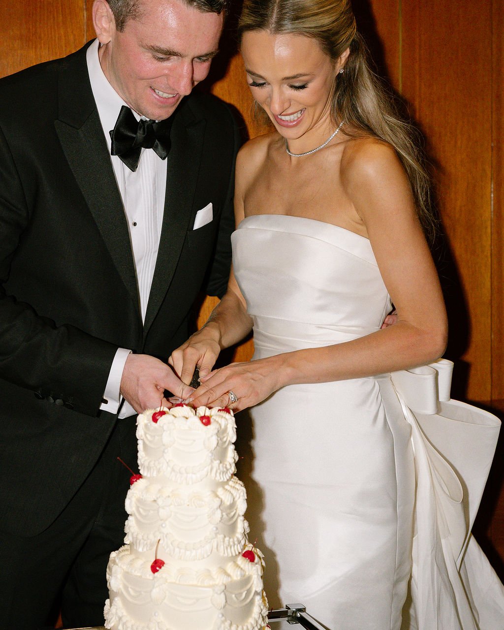 Bride and groom cutting their wedding cake together, both smiling, wearing formal wedding attire. The bride is in a strapless white wedding gown with a ribbon detail, and the groom is in a black tuxedo with a bow tie.