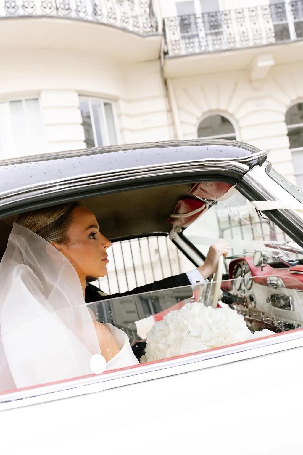 A bride with a white veil sitting in the passenger seat of a vintage car, holding a bouquet of white flowers, with a city building in the background.