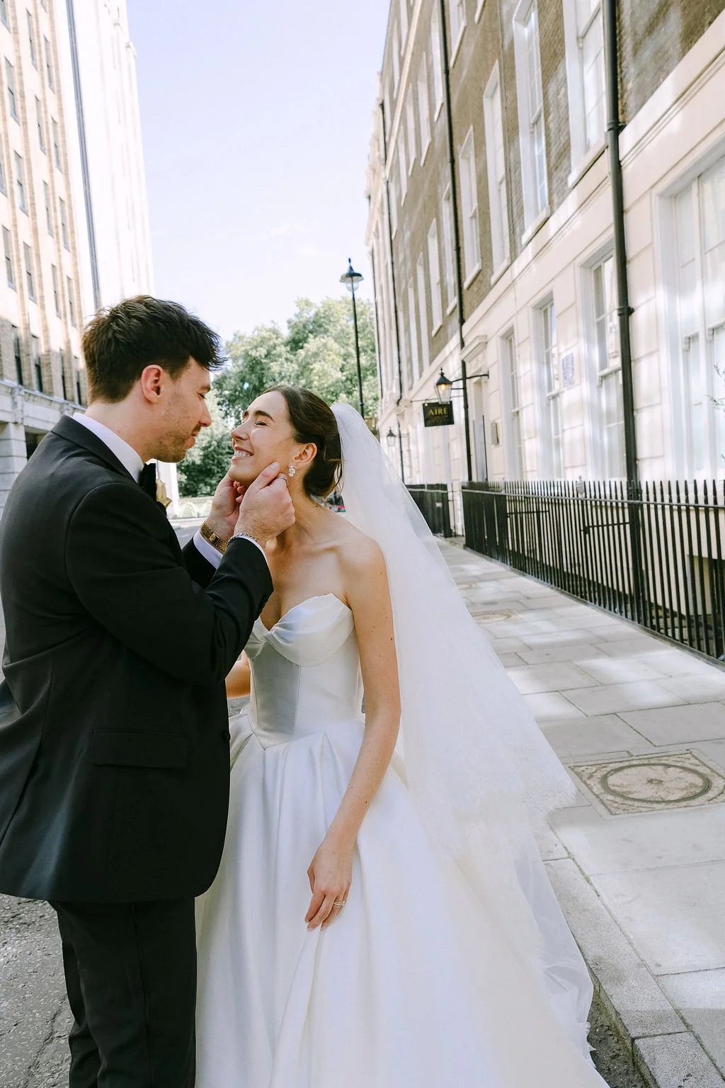 A bride and groom sharing a moment on a city sidewalk, with tall buildings and streetlamps in the background.