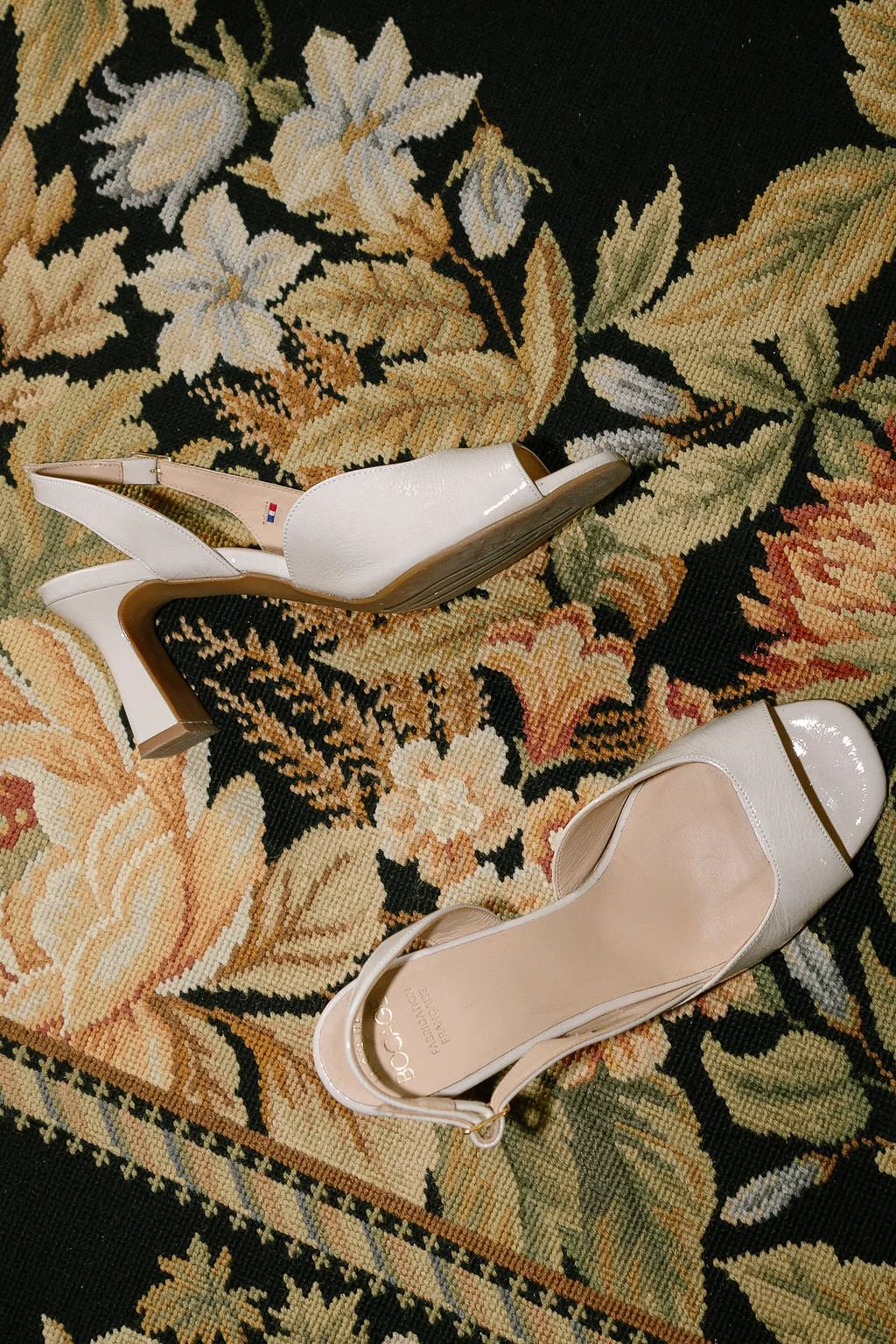 A pair of white high-heeled shoes with open toes resting on a floral-patterned rug.