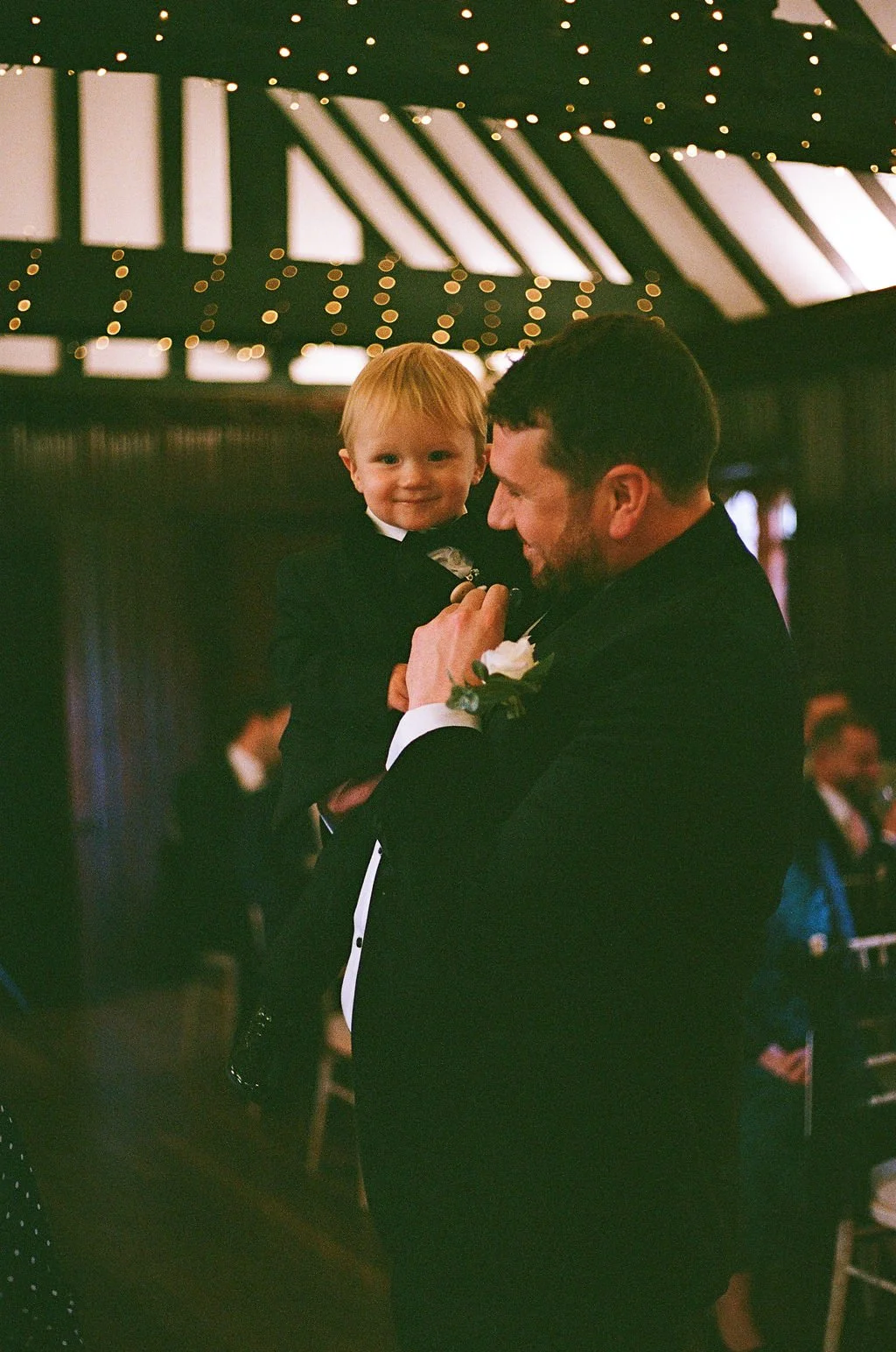 A man in a tuxedo holding a young boy, both smiling at a wedding reception with hanging lights and other guests in the background.