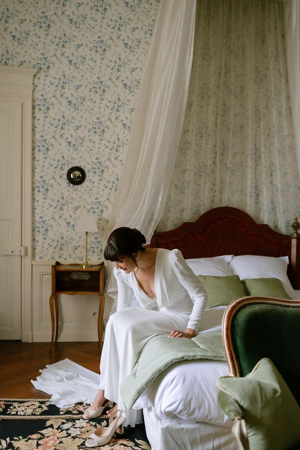 A woman in a white dress is sitting on a bed in a cozy, vintage-style bedroom, adjusting her shoes.