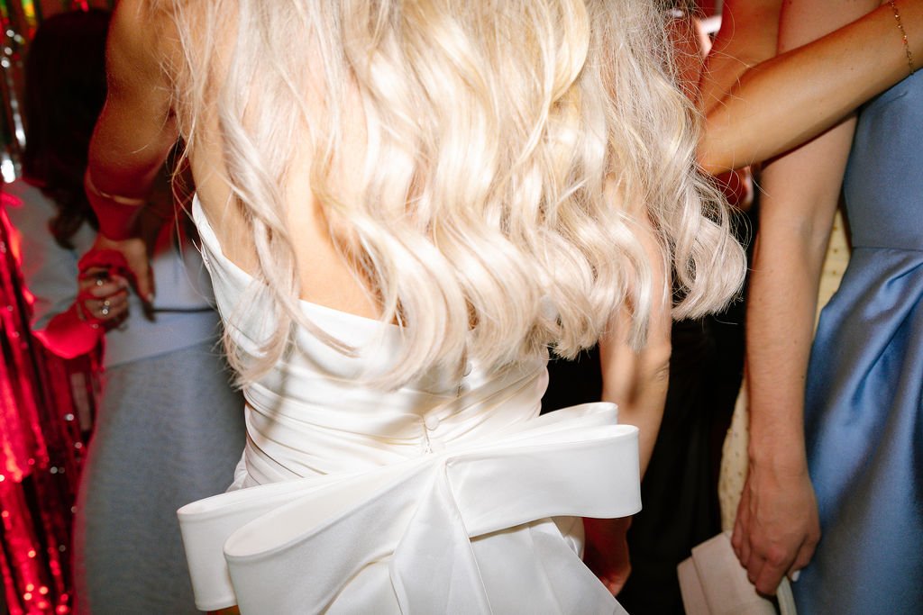 Close-up of a woman wearing a white dress with a large bow at the waist and long, wavy blonde hair, at a social event or party.