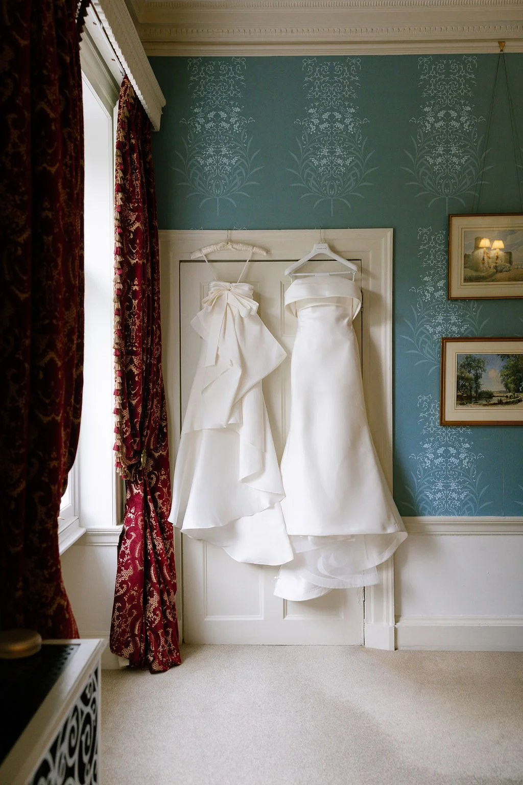 Two white wedding dresses hanging on hangers on a door, set against a blue wall with white floral patterns, near a window with red patterned curtains.