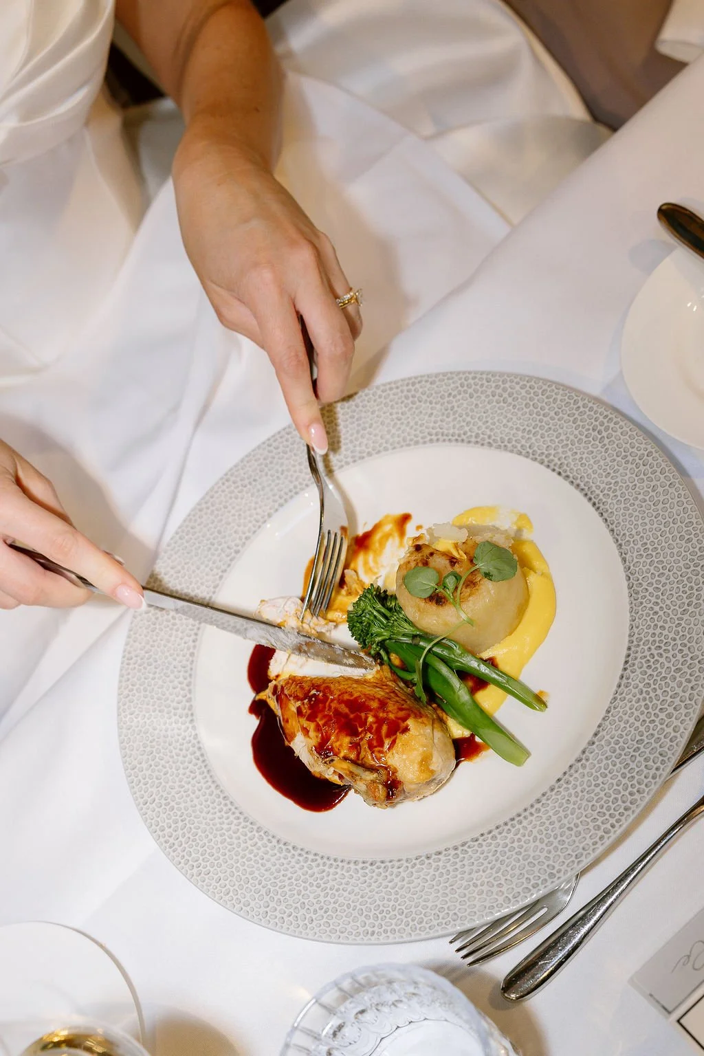 A person cutting into a plated dish of fancy food with a fork and knife, featuring green beans, mashed potatoes, a piece of meat with sauce, and garnishes.