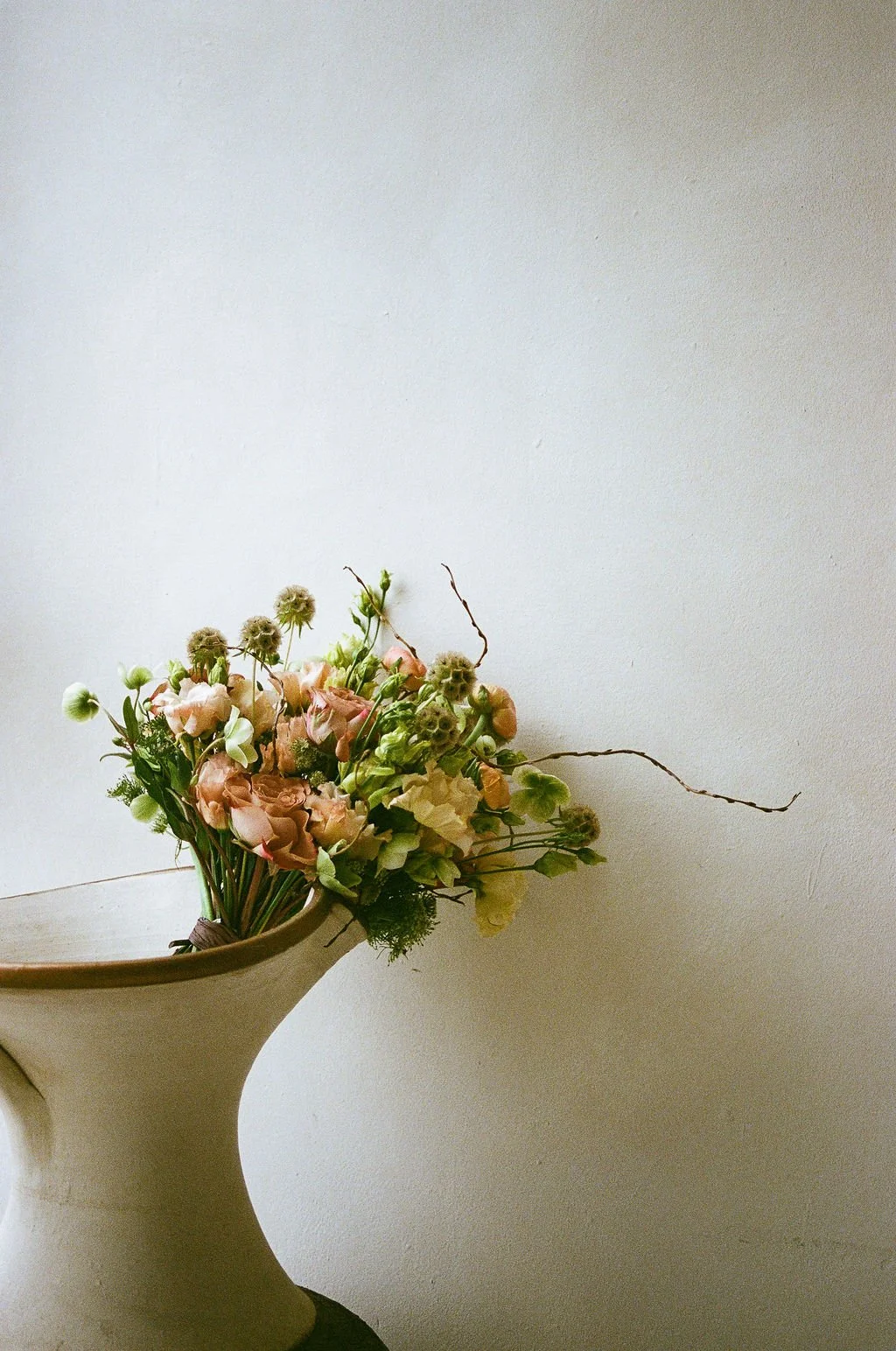 A floral arrangement in a beige ceramic vase featuring pink, white, and green flowers against a plain, light-colored wall.