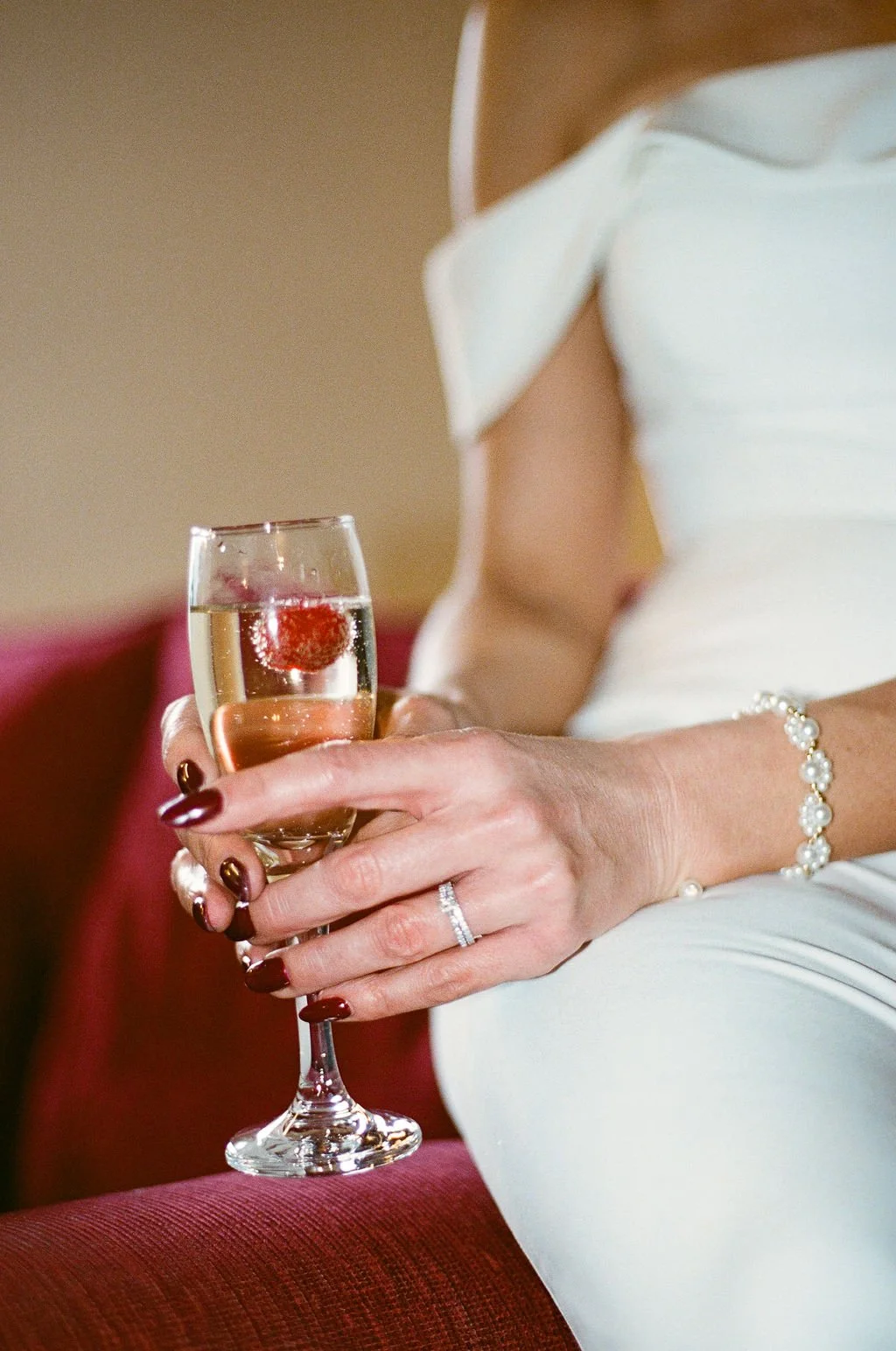 A woman in a white dress holding a glass of champagne with a cherry, wearing a pearl bracelet and a diamond ring, sitting on a red velvet chair.