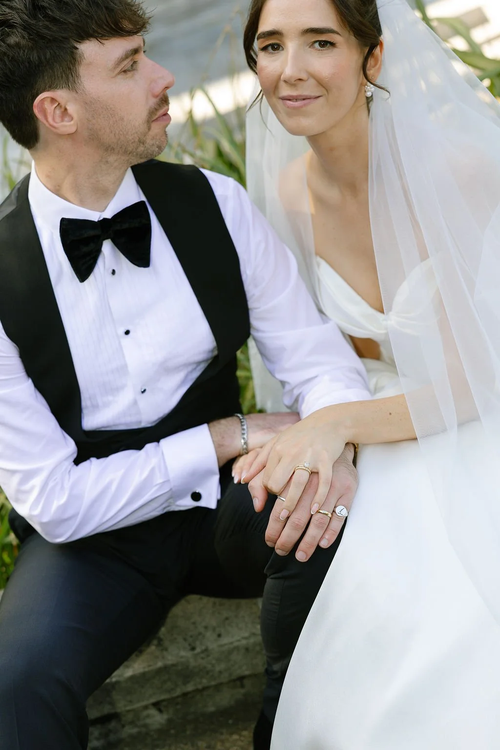 A bride and groom sitting outdoors, holding hands, with the bride wearing a white wedding dress and veil, and the groom in a tuxedo with a bow tie, looking at each other.