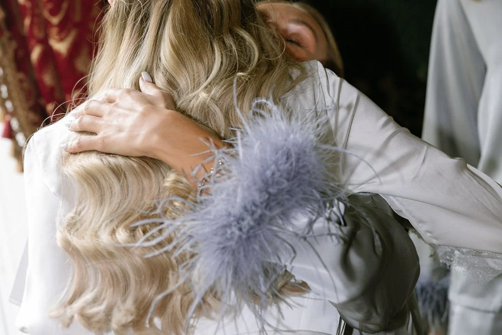 Two women share a hug, one with long blonde hair wearing a white silk robe with a feathered sleeve. The scene appears emotional and warm.