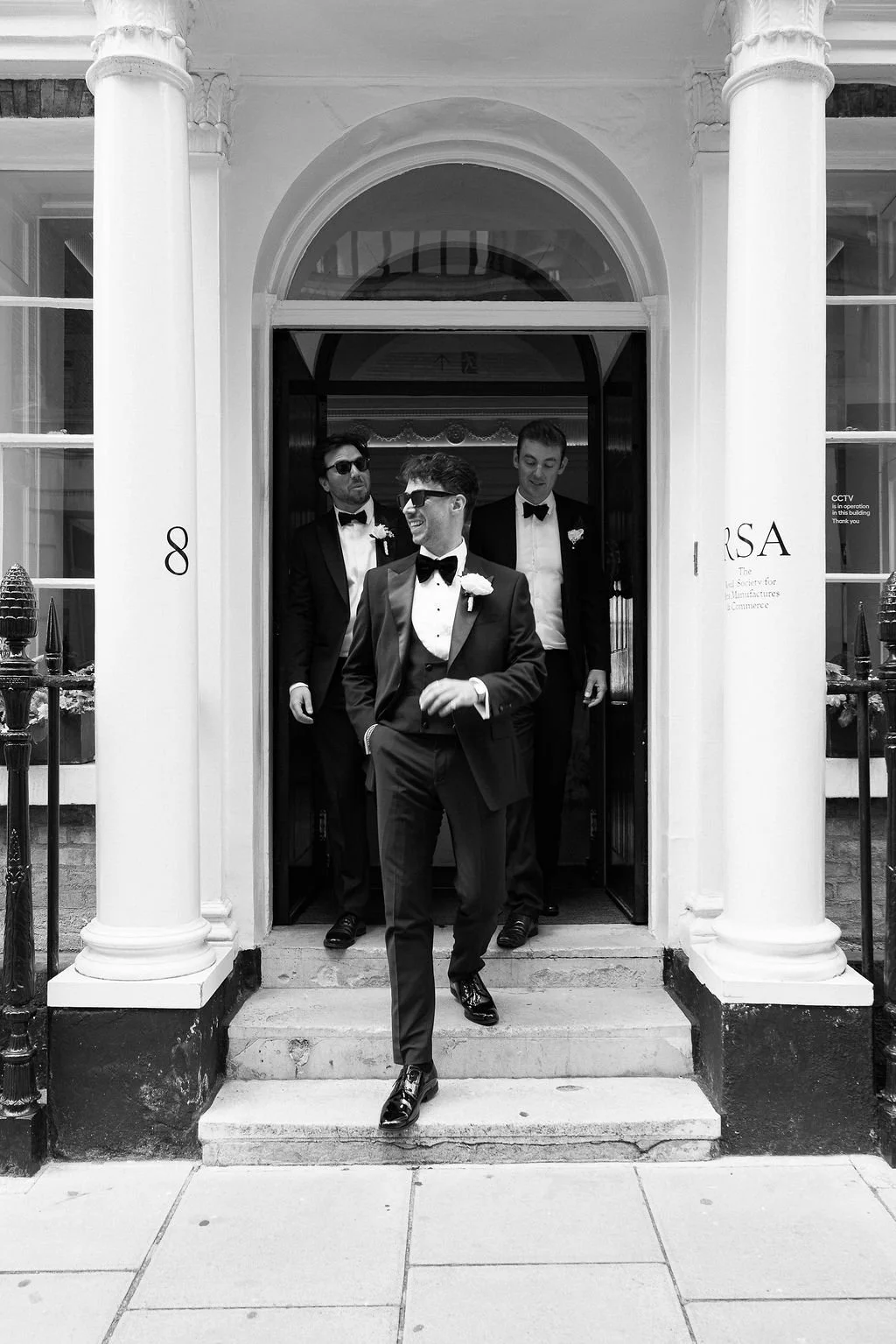 A group of three men in tuxedos with bow ties, exiting a building through a doorway. They are wearing boutonnieres, and the scene appears to be festive or formal.