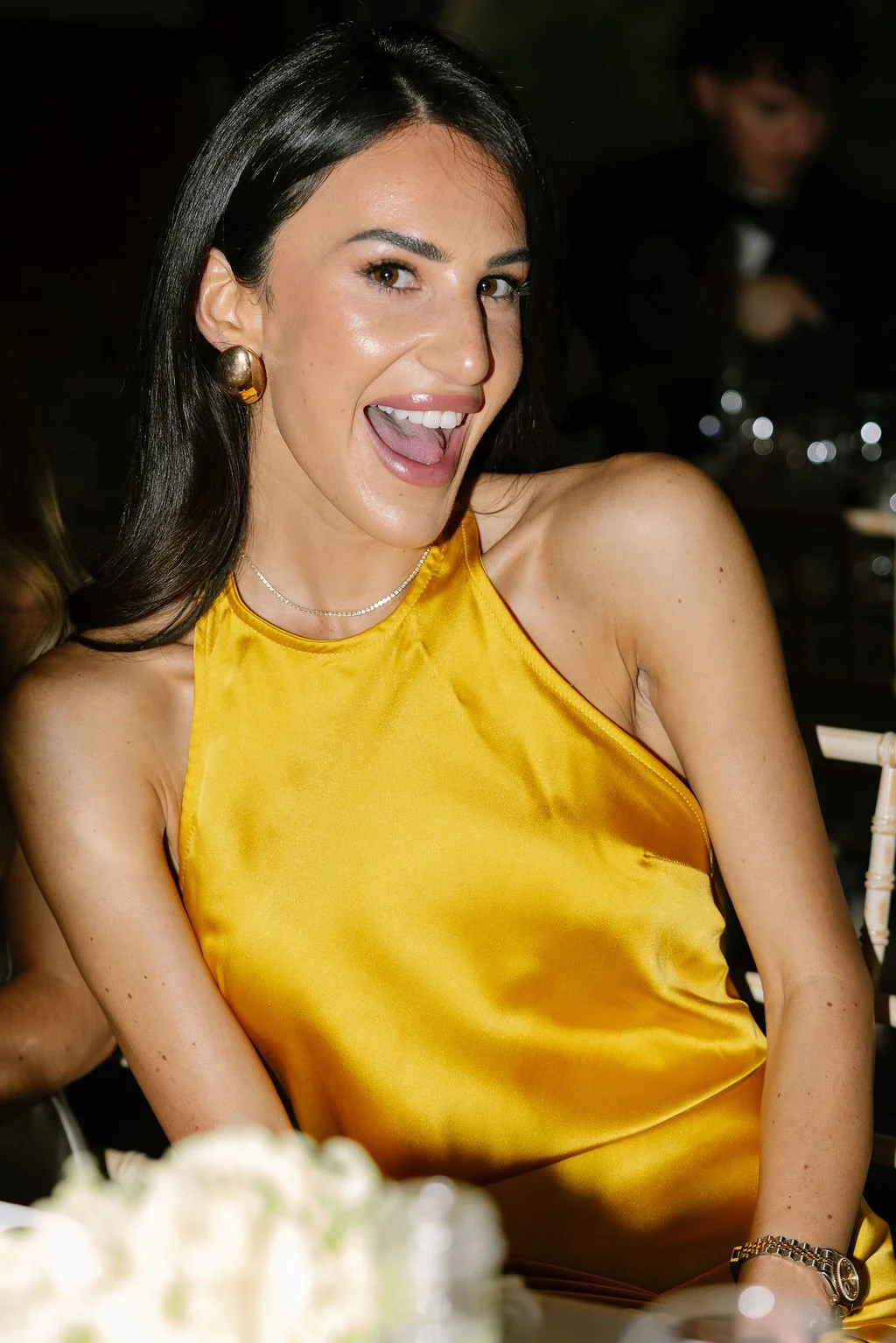A woman with dark brown hair and light skin is smiling and wearing a yellow satin sleeveless dress, large gold earrings, a silver necklace, and a watch, at a formal or celebratory event.