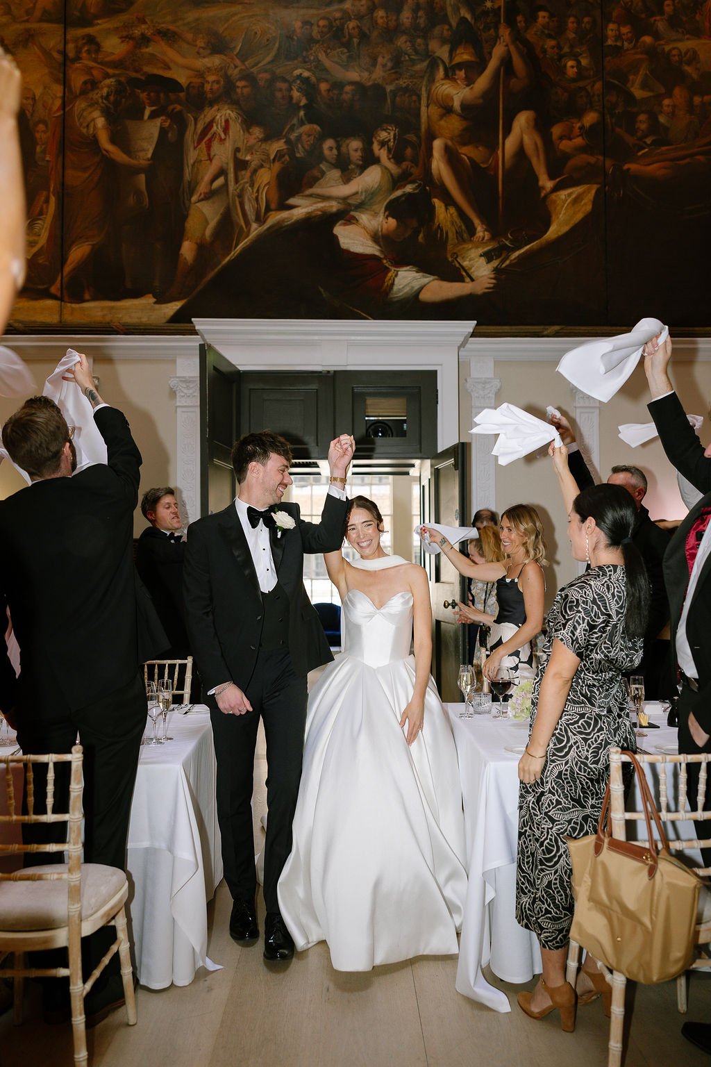 A newlywed couple celebrating with friends at their wedding reception, with guests raising napkins and smiling. A large classical painting is visible above the doorway.