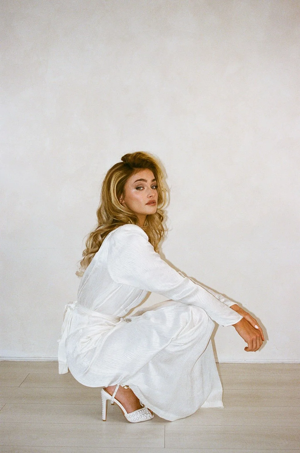 A woman with blonde hair wearing a white outfit and high heels squatting on the floor against a plain white wall.