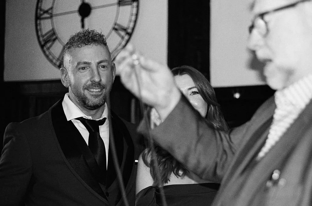 A man in a tuxedo at an event, smiling and looking to his left, with a woman partially visible behind him. An older man in glasses, also in a suit, is in the foreground, slightly out of focus.