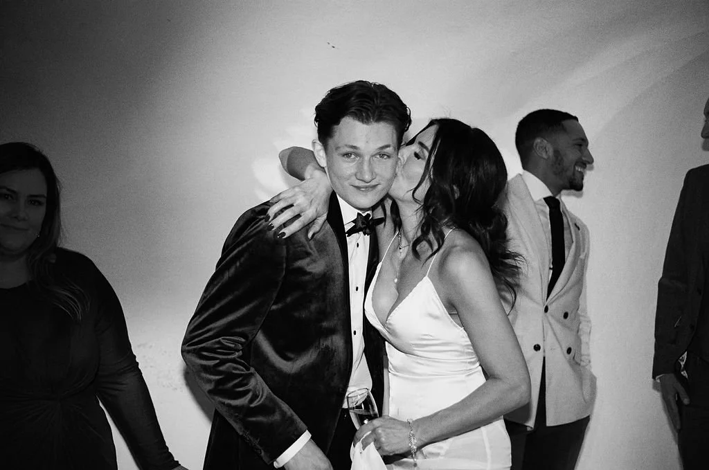 Group of people at a formal event, with a woman kissing a young man on the cheek, all dressed in elegant attire.