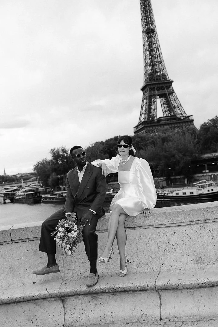 A black and white photo of a man and woman sitting on a low wall near the Seine River with the Eiffel Tower in the background. The woman is wearing a white dress, high heels, and sunglasses, and the man is in a suit holding a bouquet of flowers.
