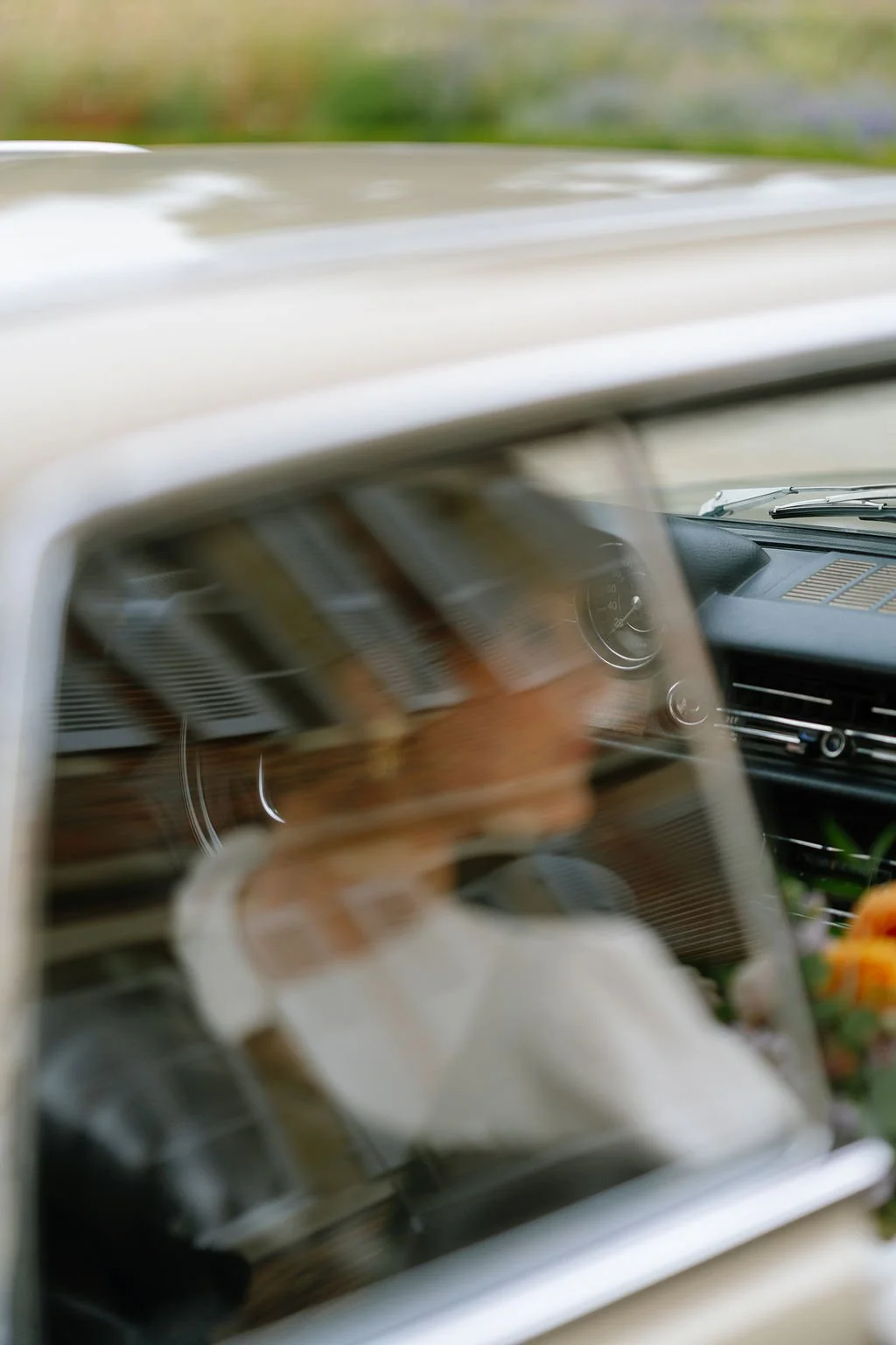 Blurred photo of a person inside a car with a reflective dashboard and steering wheel, viewed through the windshield, with some greenery outside.