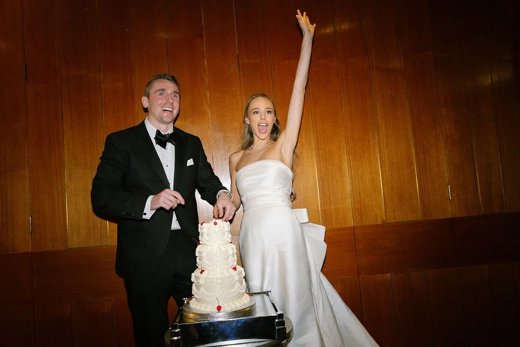 A bride and groom in wedding attire celebrating with a multi-tiered wedding cake, with the bride raising her hand and both smiling, standing in front of a wooden wall.