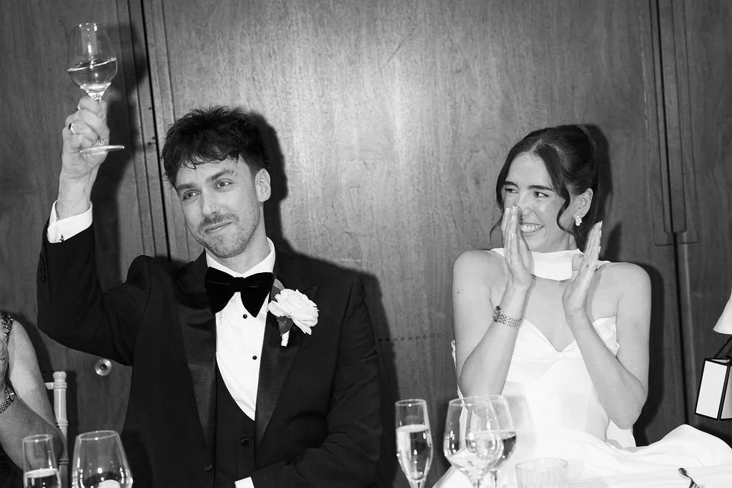 A man in a tuxedo raising a glass at a formal event, with a woman in a white dress smiling and clapping beside him.