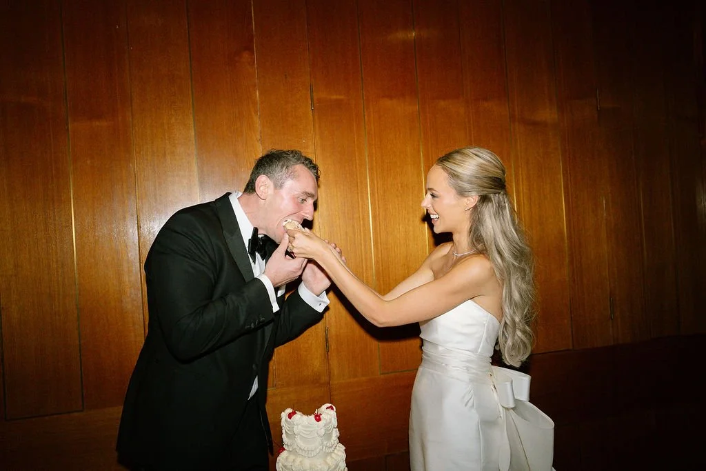 A bride and groom at their wedding reception, with the groom eating wedding cake and the bride holding his hand, smiling at each other.